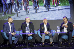 Czterech mężczyzn siedzi na fotelach ustawionych na scenie,  za nimi na dużym ekranie wyświetla się zdjęcie przedstawiające wyścig kolarski