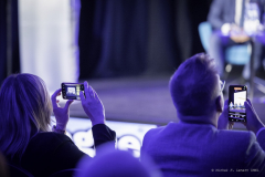 Kobieta i mężczyzna siedzą tyłem do osoby robiącej zdjęcie, oboje trzymają telefony w rękach i nagrywają trwającą konferencję