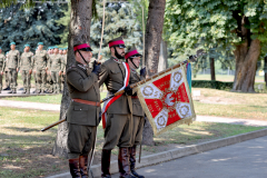 Trzech ułanów stoi ze sztandarem na placu