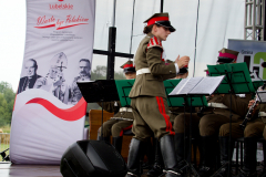 Dyrygentka w mundurze stoi na scenie przed orkiestrą. Za nią widać baner z napisem Warto być Polakiem