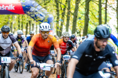 Skupieni rowerzyście jadący trasą otoczoną drzewami.