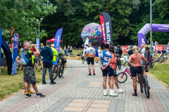 Grupa ludzi, niektórzy z rowerami, stojących na chodniku i obserwujących wydarzenie.