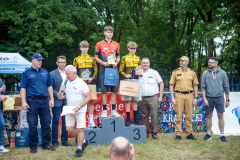 Zdjęcie przedstawia podium oraz trzech młodych mężczyzn, rowerzystów, stojących z nagrodami.