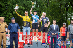 Zdjęcie przedstawia trójkę ludzi na podium unoszących wygrane puchary w górę.