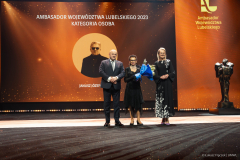 Marszałek WL i posłanka na Sejm RP stoją na scenie i pozują wspólnie do zdjęcią z mamą laureata, która odbiera w jego imieniu statuetkę