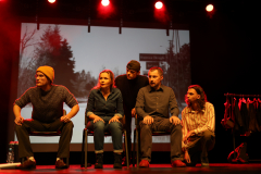Czterech aktorów i jedna aktorka podczas występu, siedzą na krzesłach ustawionych na scenie i patrzą wszyscy w jednym kierunku