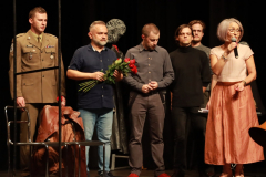 Grupa aktorów stoi na scenie po zakończeniu przedstawienia, Konferansjerka wypowiada się do mikrofonu, jeden z aktorów trzyma kwiaty  w ręce