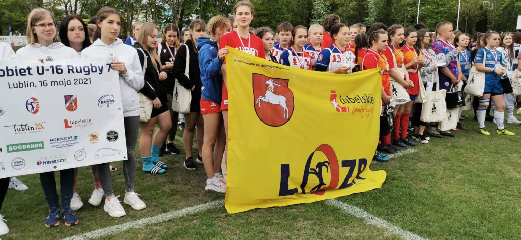 drużyny kobiece rugby na otwarciu zawodów trzymające flagi z logotypami klubów i sponsorów