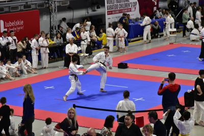 Zawody w judo. Na macie walczy dwóch przeciwników, dookoła nich znajdują się widzowie i pozostali uczestnicy zawodów, łącznie na zdjęciu widać około pięćdziesięciu osób