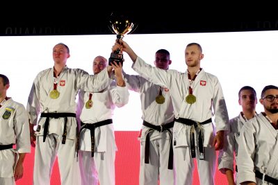 Czterech złotych medalistów w zawodach karate, stoi obok siebie i wspólnie trzymają zdobyty puchar. każdy z nich ma na szyi powieszony złoty medal, wszyscy są ubrani w kimona sportowe. Obok nich znajduje się kilku innych zawodników